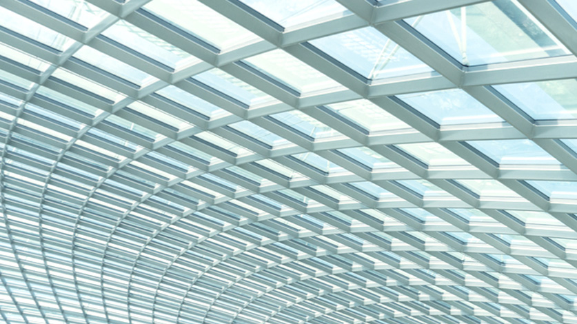 Curved glass roof with a white metal framework in a modern architectural design, showcasing a pattern of parallel lines and light flooding through the translucent panels