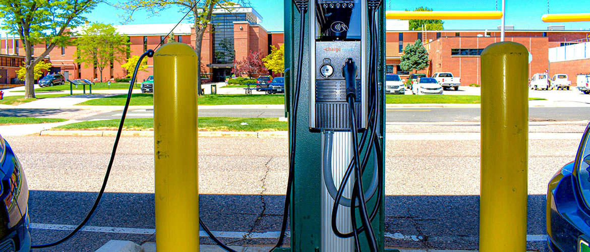 Close-up of two electric vehicle charging ports between two cars, with yellow posts and a red brick building in the background