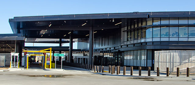 Cropped view of two-level steel and glass building, with covered driveway lanes, yellow traffic poles and a row of short red bollards surrounding the building