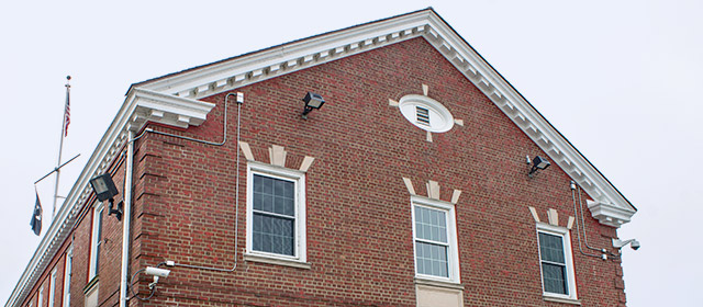 Close up of red brick building facade with three rectangular windows and an oval vent above the center window, with U.S. flag in back at left