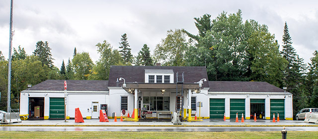 White one-level building with six garage spaces, orange traffic cones and a covered drive-through, with trees in the background and green grass in front