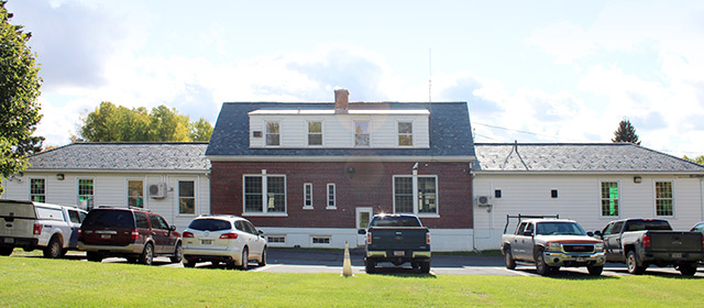 One-to-two story three-part white and red brick building, with six vehicles parked along it, and green grass