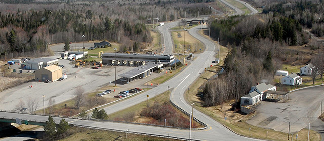 Aerial view of several low silver, white and tan buildings, with traffic check lanes, highways, parking lots, and grasses and forests around