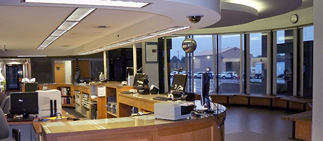 Office interior view with curved, blond-wood desk area, white ceilings and a view out of a window bay to see a tan building and vehicles outside in the distance