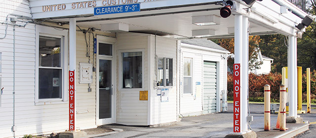 Cropped close up view of a white shingled building entrance, with cover over one driving lane, yellow posts, and signage that says United States Customs in gold, Clearance 9'-3" in blue and Do Not Enter twice in red