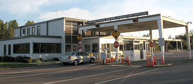 Quarter view of the front of a low white building with two booths next to two lanes, and one gold car entering, and a cover over the bays with the sign United States Border Inspection Station across it