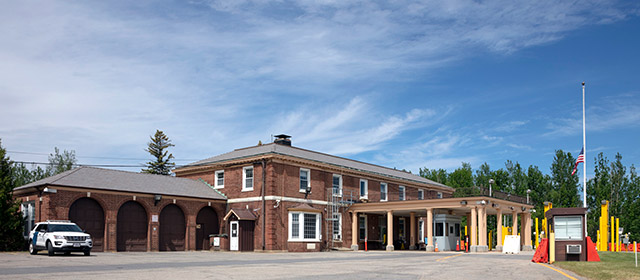 Two-level red brick building with a canopy in front with columns, yellow traffic guards, an official vehicle parked on the left, and a blue sky and trees in the background
