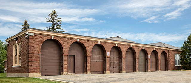Angled view of a red brick building with tan trim, and eight arched garage door entrances, with a concrete driveway in front, several trees in the background and a blue sky with clouds