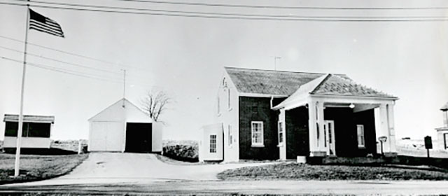 Street-level black and white view of a gabled-roof building on the right, with a canopy over one lane in front of it, and a white garage building to the left of it, and another small building and a U.S. flag on a pole on the far left