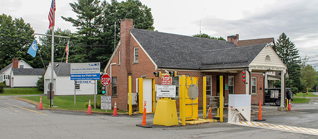 Low red brick building with gabled roof, chimney and canopy that extends over one lane outside of the entrance door, with yellow guards and stop signs, orange traffic cones and three white buildings on the left in the distance