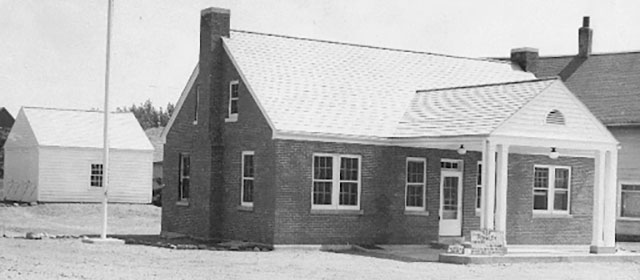 Black and white photo of a brick building with a chimney and dormer roof and a canopy outside of the door over one driving lane, with a flag pole and a white building on the left