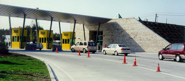 Angled street-level view of a curved awning that extends from a tan brick building entrance over three paved lanes with yellow booths beside each lane, orange traffic cones and four cars lined up in one lane