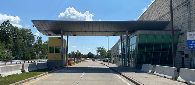 Driver's view of a curved canopy that extends over two paved lanes, with a yellow booth at the left and trees along the left side, and to the right, the angled, glass entrance to a two-level, tan-brick building