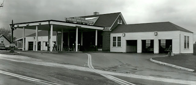 Dramatic black and white street-level view of a white building with a canopy over two lanes in the center front entrance, with three garage bays on either side, and a cloudy sky above