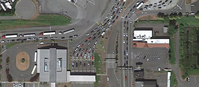 Aerial view of a border crossing station, with cars and trucks in line in paved lanes, with buildings and grassy areas nearby