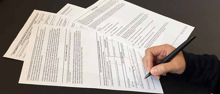 Several papers and forms with a hand signing one of them with a pen