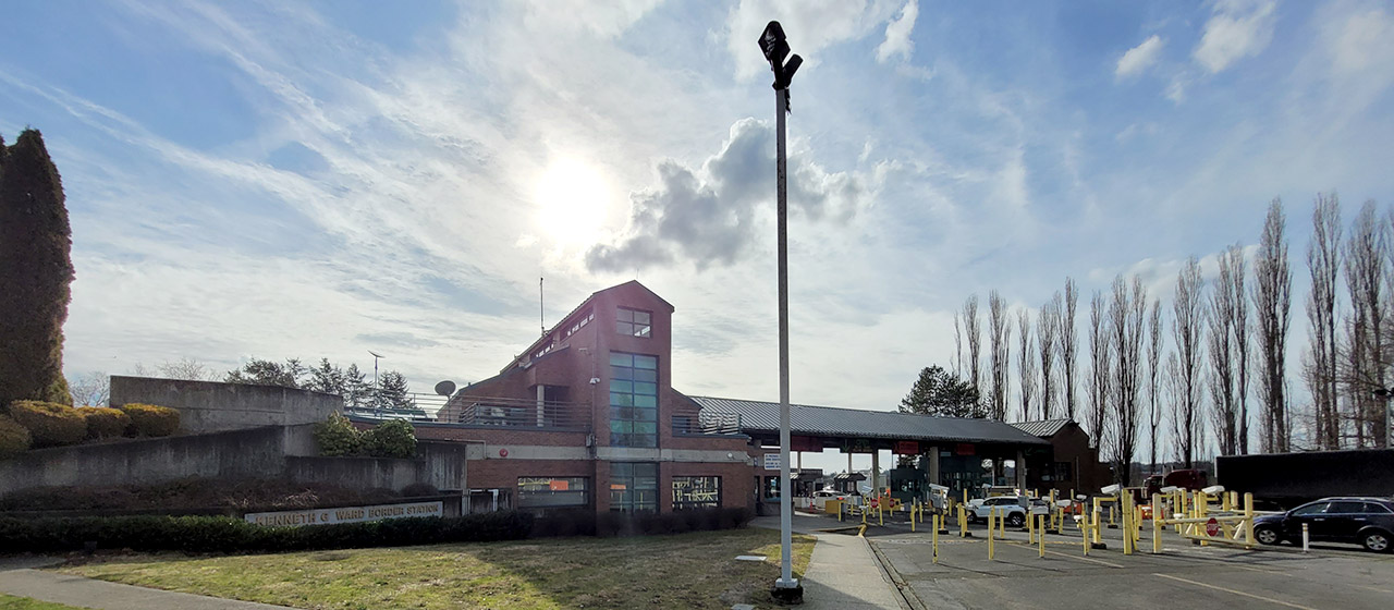 Dramatic backlit photo of a multi-level red brick building, with a canopy over several traffic lanes on the right, and trees flanking each side, with a lightpost in the foreground center