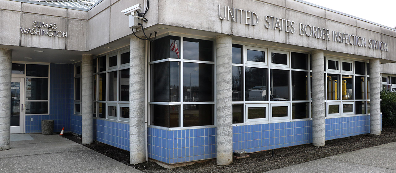 Cropped view of the front of a 1-level cement building with small columns, blue tile running under the windows, with words Sumas Washington and United States Border Inspection Station along the top near the roof