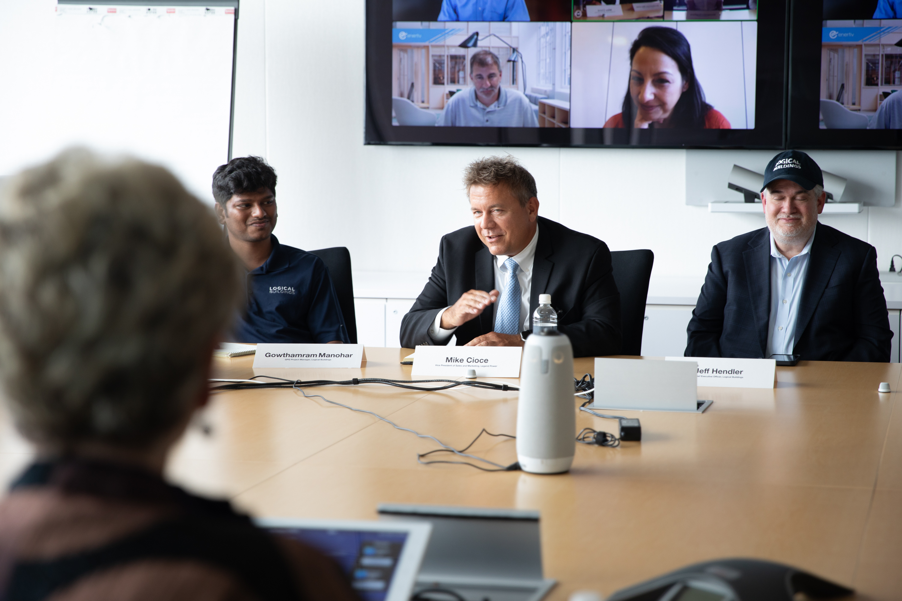 Three men sitting at a table having a discussion with other people on a screen behind them