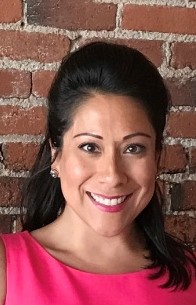 A person wearing a pink shirt, smiling, in front of a brick wall.