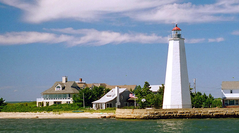 A white lighthouse on the right next to a body of water with homes on the left.