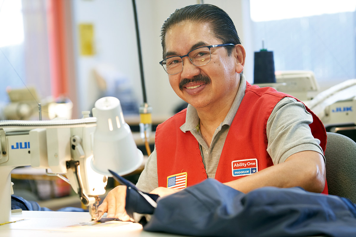 Smiling Asian male employee with a moustache and wearing a red vest sitting at a sewing machine