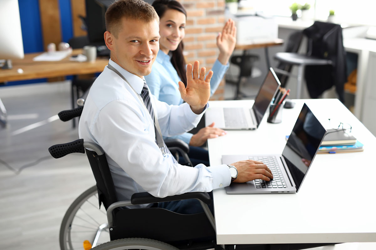 Two people in wheelchairs at a desk with laptops looking at camera and waving