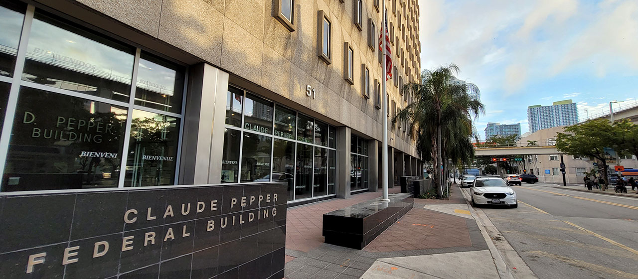 Light stone multi-story cube-shaped building with signage saying Claude Pepper Federal Building, with palm trees in front