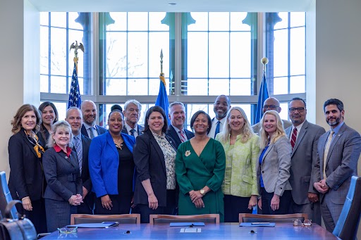 Sixteen people stand behind a table posing for a photo with three flags behind them.