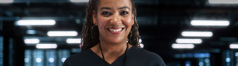 Smiling Black woman with hoop earrings and black v-neck sweater in front of a wall of servers