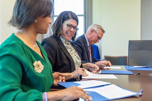 Three people sit at a table with blue folders in front of them. They are each signing and passing the folders to the next person