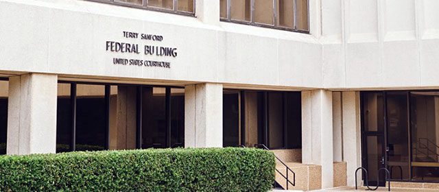 Entrance to a light stone building in the Brutalist style, with a green hedge and signage saying Terry Sanford Federal Building