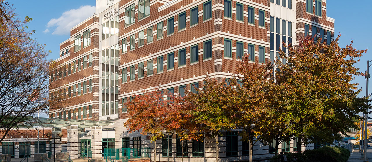 Angled view of a red multi-story brick building with white decorative stripes, with fall trees in the foreground
