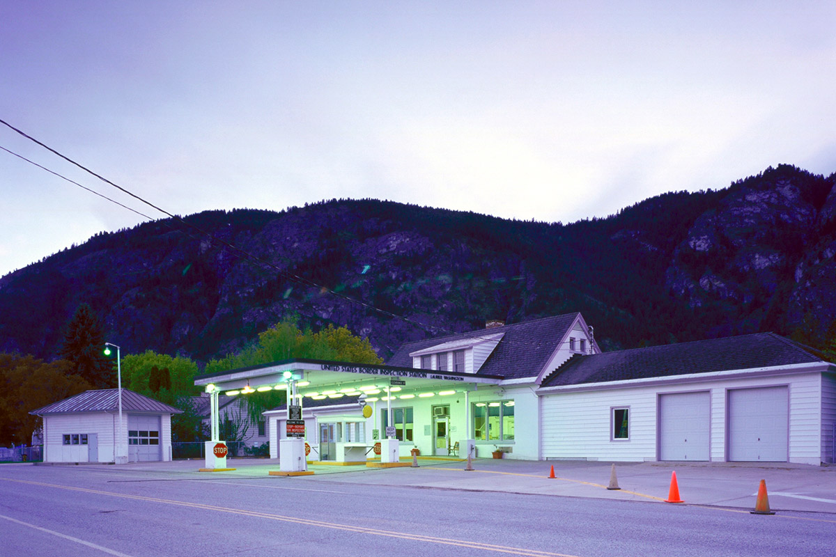 A white mostly one-level check-station building with green lighting and garage at dusk or dawn in front of a range of hills with trees