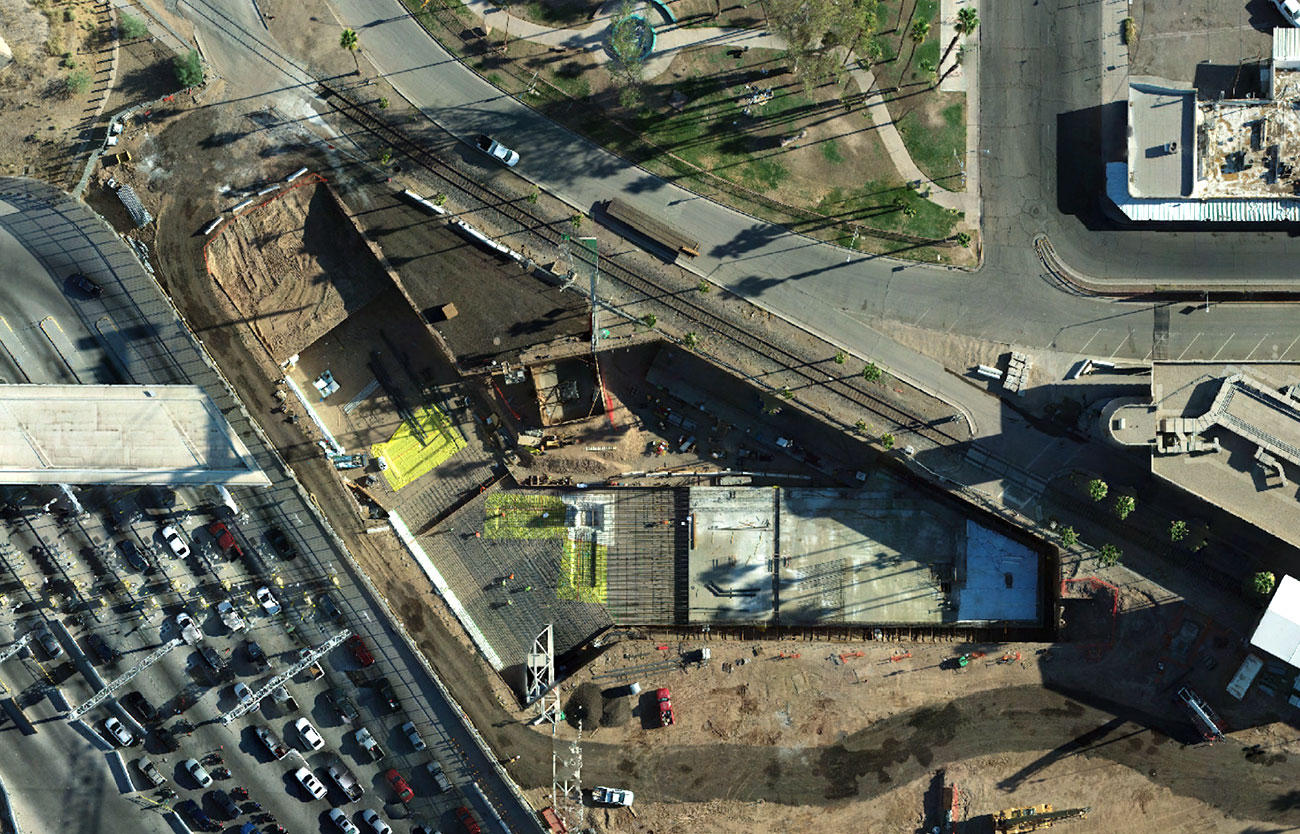 Overhead view of a large building construction area, with several lanes of cars to the left/west of it