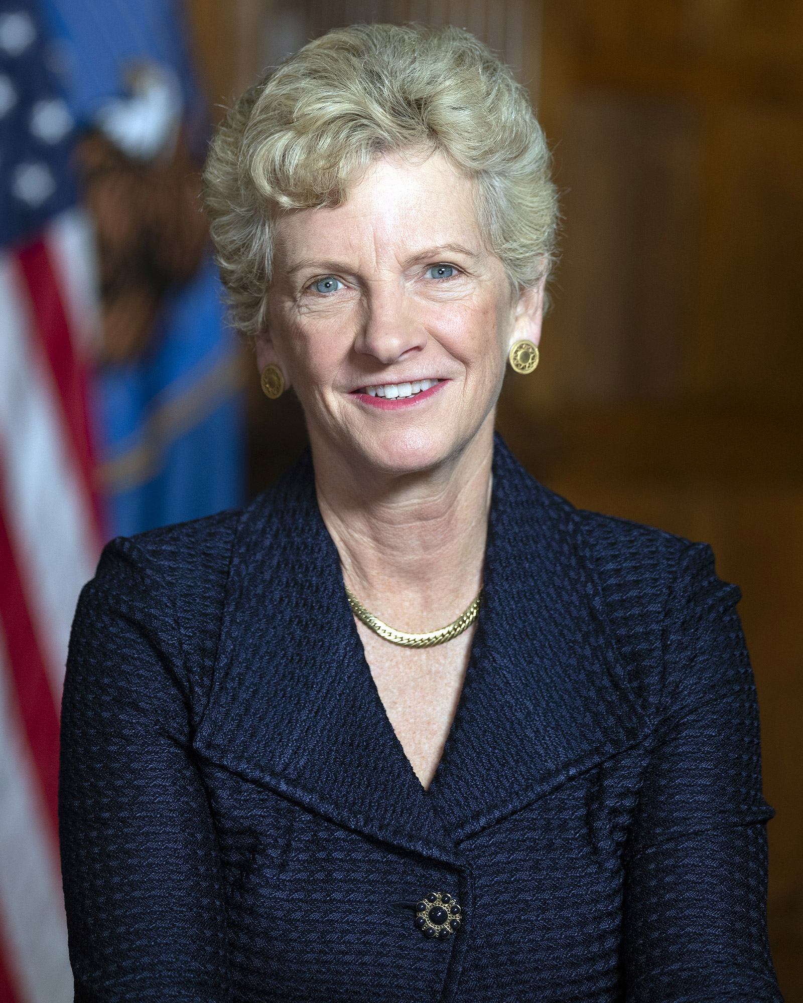 Smiling woman with short blond hair, wearing a navy jacket, and gold necklace and earrings