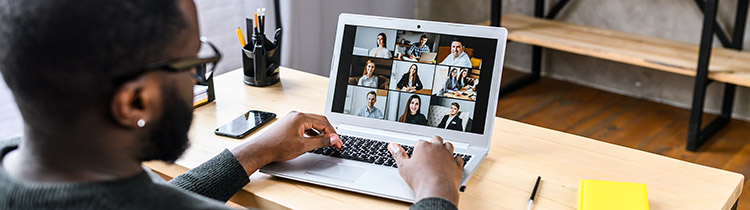 Looking over a young man's shoulder at a virtual meeting on a laptop on the desk in front of him