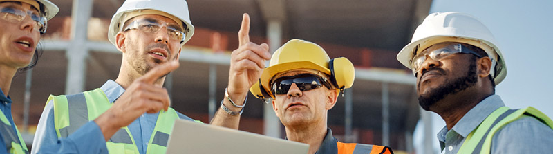 Four people in construction hats and safety vests talking and pointing