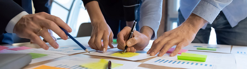 Close up of table top with three people pointing and writing on sticky notes on printed graphs