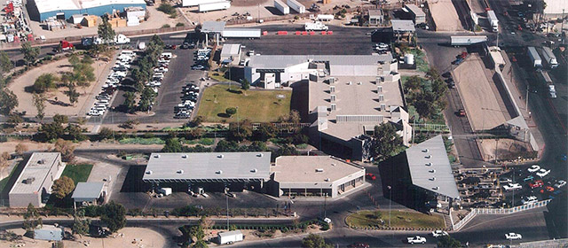 Aerial photo of a group of buildings with roads and green areas around, with lines of cars in five lanes lined up to go through inspection lanes