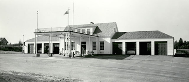 Black and white street-level view of a low white building with a canopy over three lanes in the center front entrance