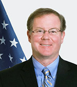 Portrait of a smiling man with glasses and brown hair in a black suit with blue shirt and tie in front of a U.S. flag