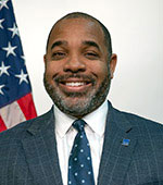 Portrait of a smiling Black man in a navy suit and tie with an American flag in the background