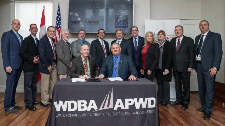 Wide shot of two GSA and Windsor-Detroit Bridge Authority officers posing seated at table with signed sublease for the U.S. land port of entry at the Gordie Howe International Crossing with team members standing behind.the ceremony Nov. 13, 2019, in Windsor, Ontario, Canada.