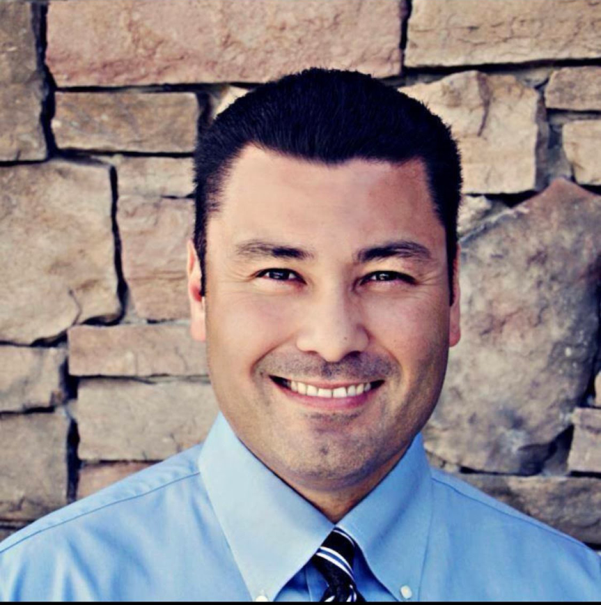 Headshot of Matt Ocana wearing a blue shirt and blue tie with yellow stripes against a brick wall