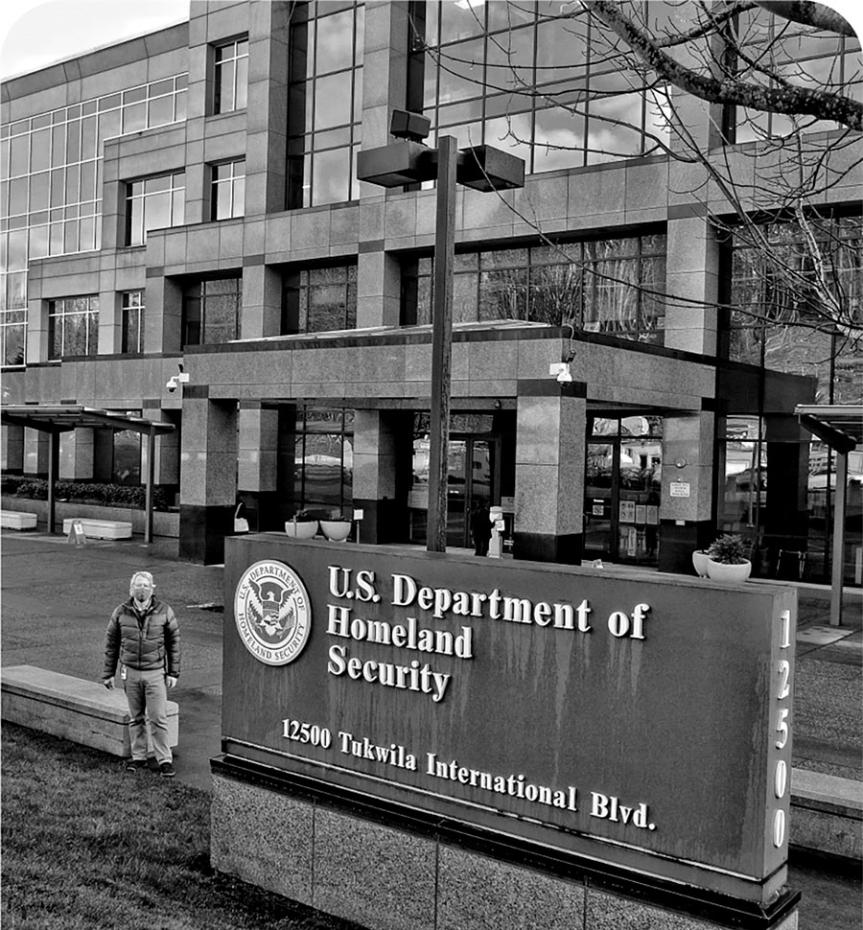 A man in a puffer jacket and facemask standing in front of a stone and glass building, next to a sign U.S. Department of Homeland Security