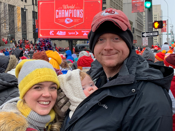 Two adults and a child, all wearing coats and hats, standing on a crowded street corner with a Kansas City Chiefs World Champions sign in the background.