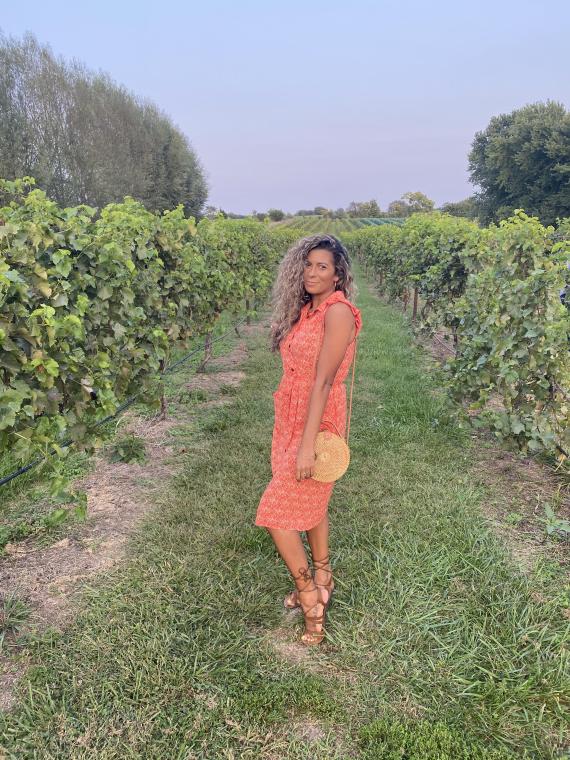 Woman with medium-colored hair wears a sundress and gladiator sandals holding a wicker purse in a vineyard.
