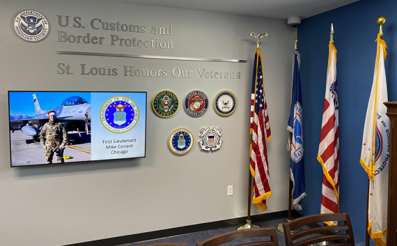An office wall with chrome lettering saying “U.S. Customs and Border Protection St. Louis Honors Our Veterans;” a monitor with photo of Veteran, rank, name, and location; large seals of DHS, Army, Marine Corps, Navy, Air Force, and Coast Guard; a U.S. flag and three others on poles; chairs and a display case in the foreground.