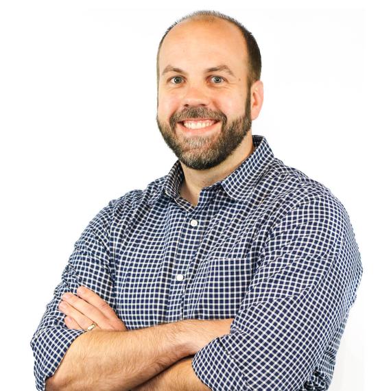 Jacob Fajen, with facial hair and arms crossed, wearing a button down shirt with top button undone and sleeves rolled to elbows